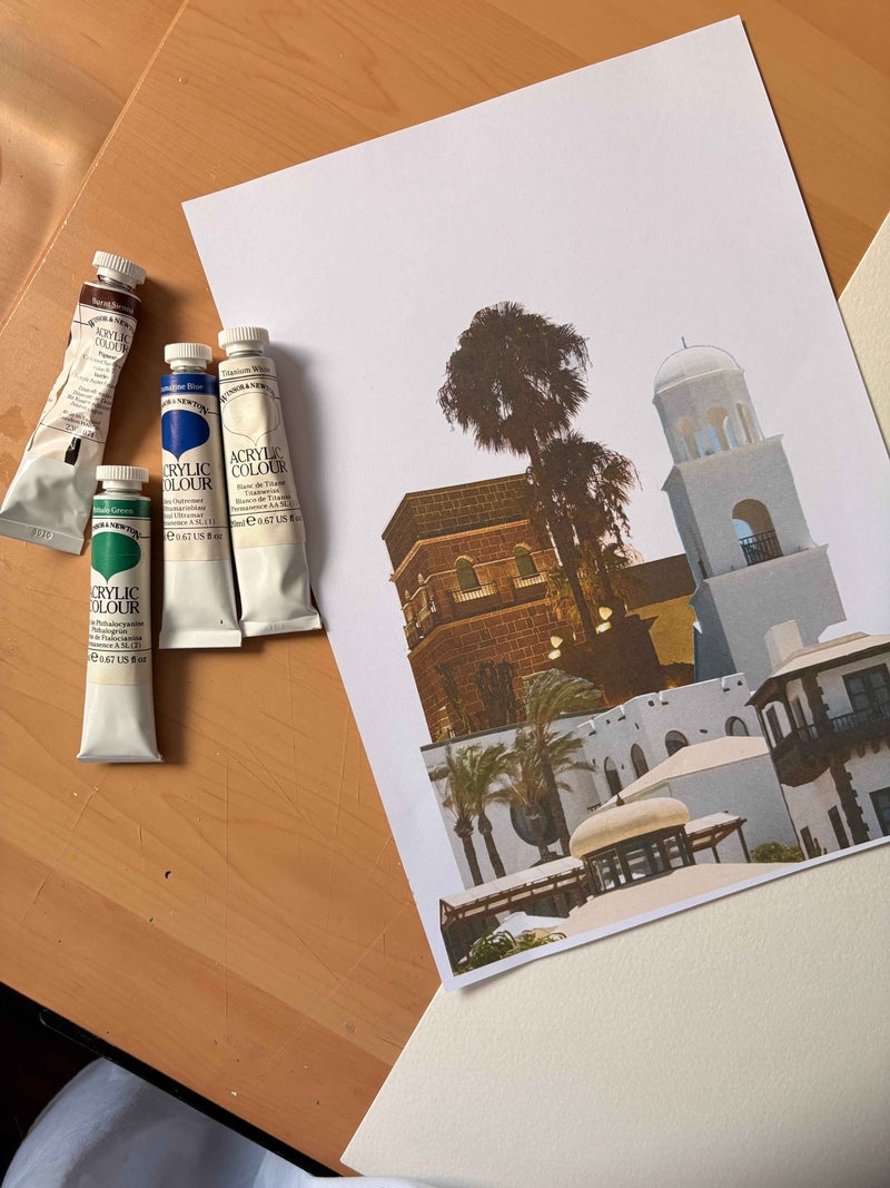 A printed art card or poster showing a Mediterranean-style building scene with a palm tree and white domed structure, placed on a wooden table next to three tubes of acrylic paint labeled Royal Poincé Color. its the image composition of Canary Islands series No.1