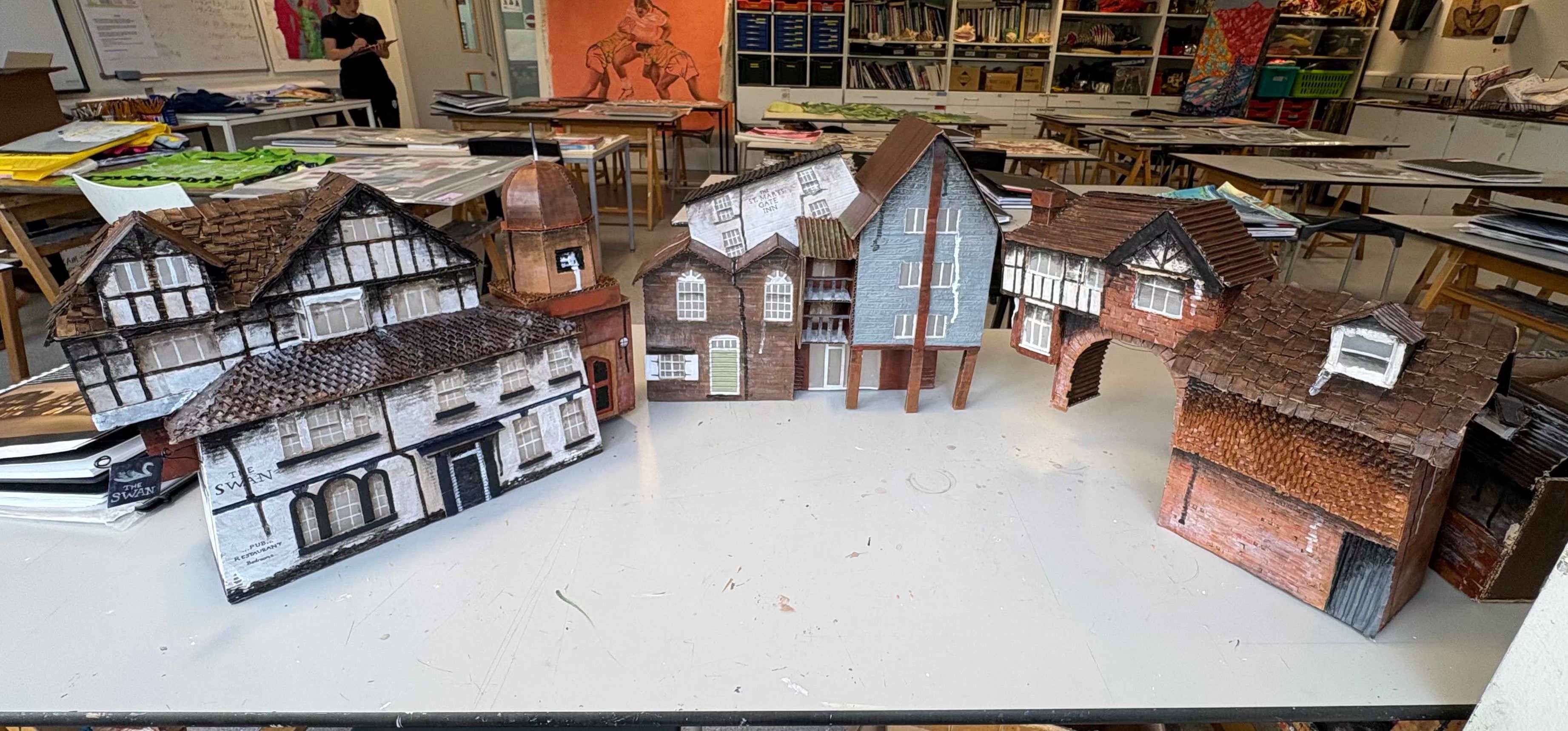 A collection of handcrafted paper model houses and buildings arranged on a white tabletop in a workshop or classroom setting.