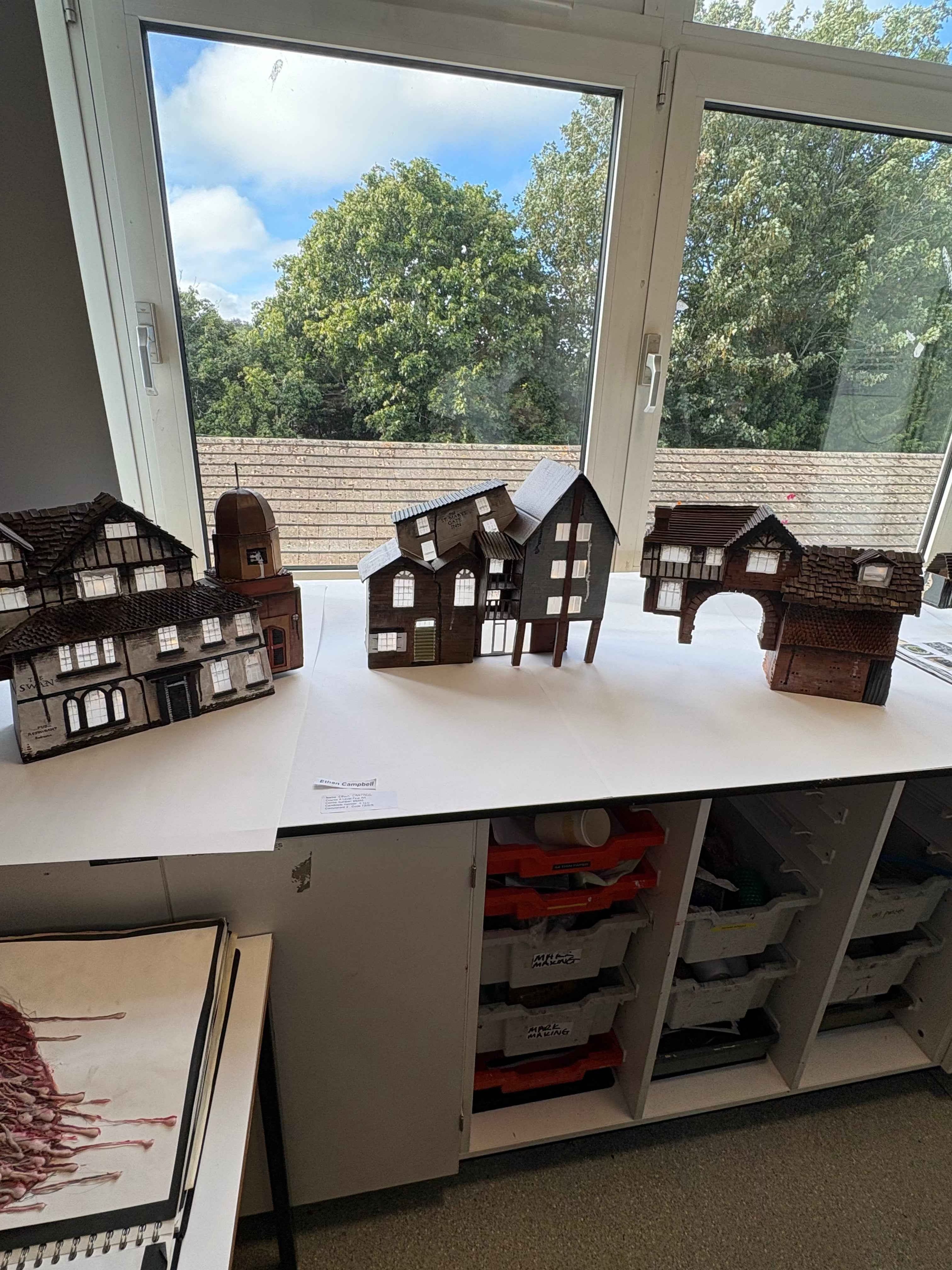 Several small wooden house models with dark brown roofs and white-framed windows are arranged on a white tabletop in front of a large window. Storage bins are visible underneath the table, and trees and sky are visible outside. This is my component 2 final piece for A-level