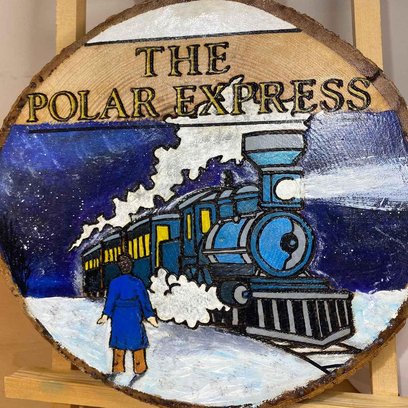 A round wooden plaque featuring a painted scene of 'THE POLAR EXPRESS' train traveling through snow, with a person in a blue coat standing beside it. The plaque is displayed on a wooden stand.