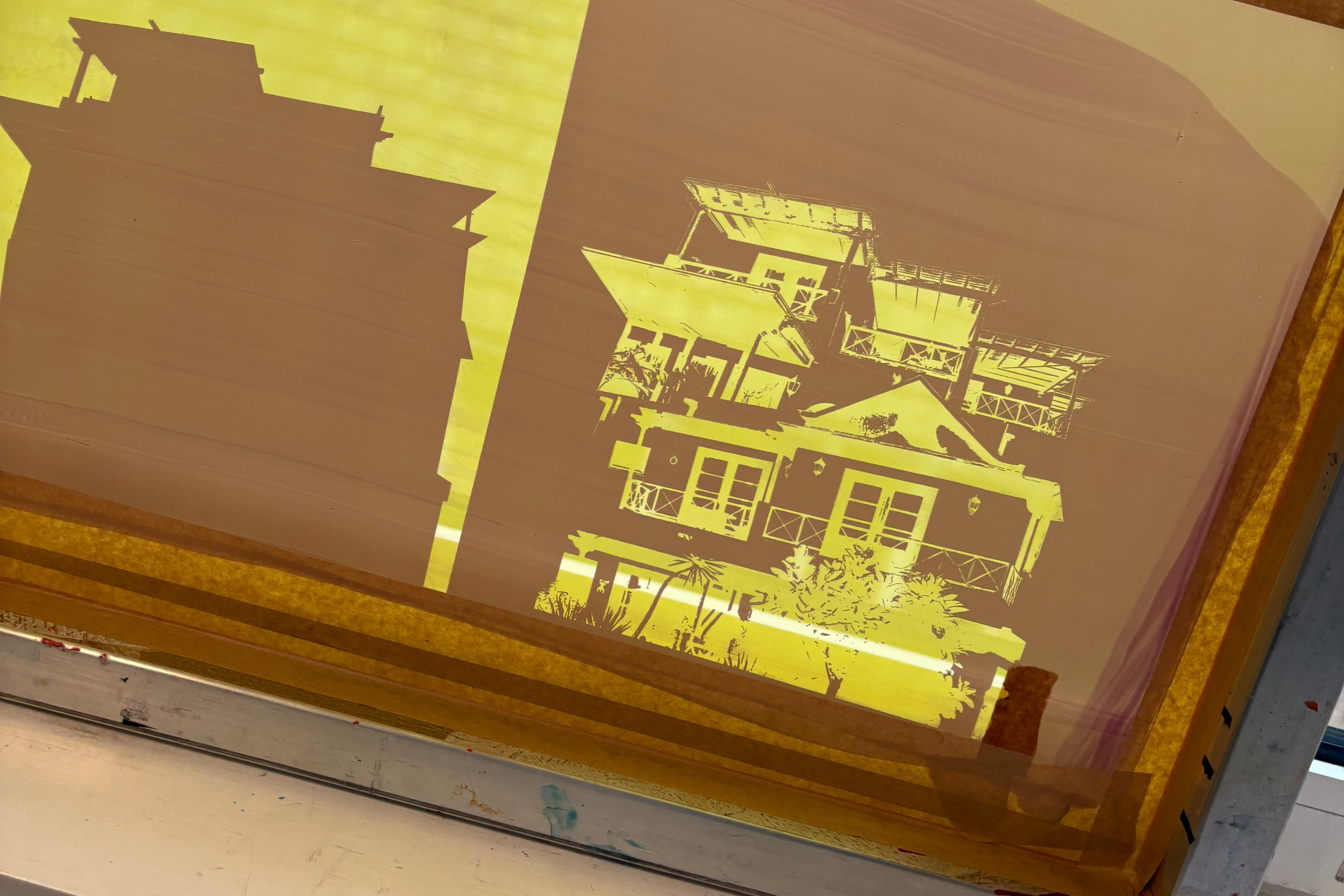 A screen-printing frame with a yellow and brown stencil design showing house silhouettes, resting on a white surface with a window and bare trees visible in the background.