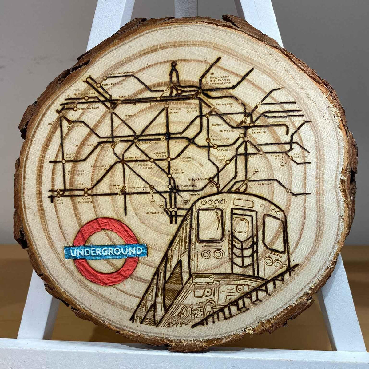A round wooden slice coaster featuring a London Underground map design with a red and blue roundel and a burnt brown train illustration, displayed on a white stand.